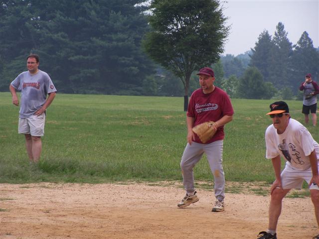 The defense (Pat and Jim G.) set as Bill stands on first with umpire Ron looking on